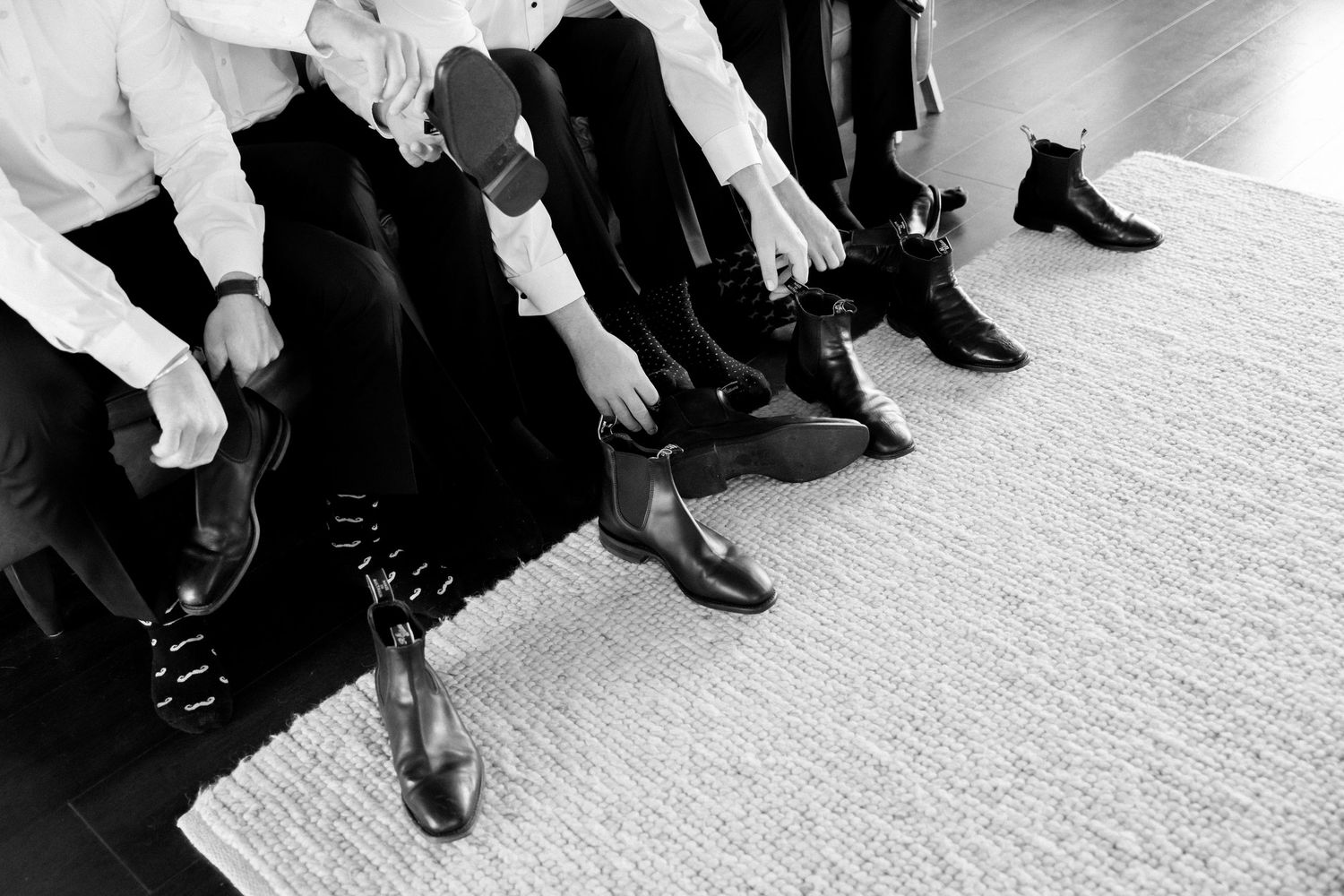 Black and white image showing people sitting in a row putting on dress shoes and formal footwear on white carpet.
