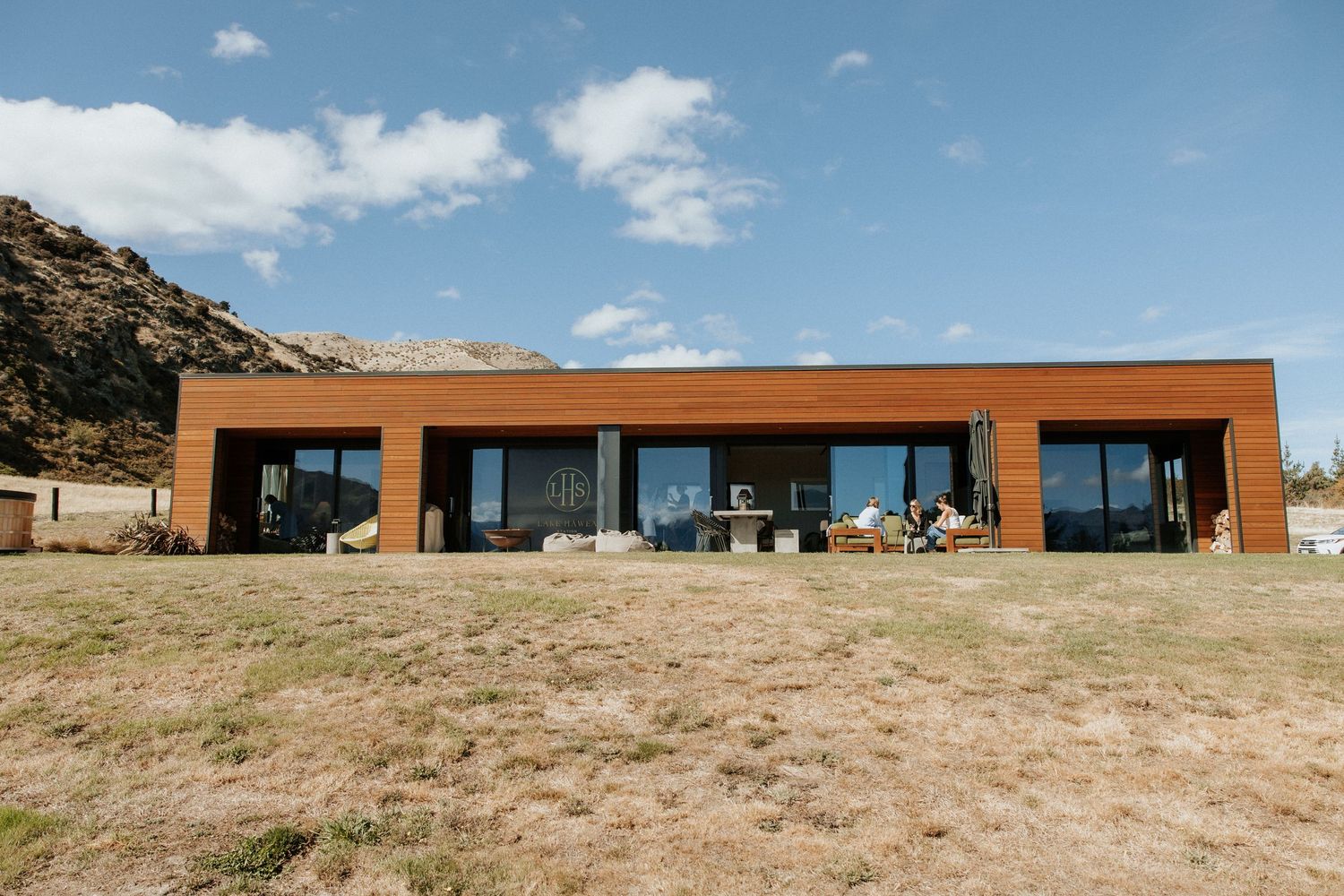 A modern rectangular building with rust-colored exterior sits nestled against a mountain landscape under blue skies.