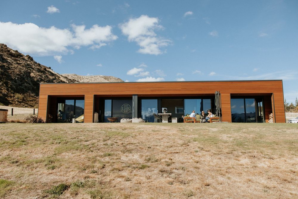 A modern rectangular building with rust-colored exterior sits nestled against a mountain landscape under blue skies.