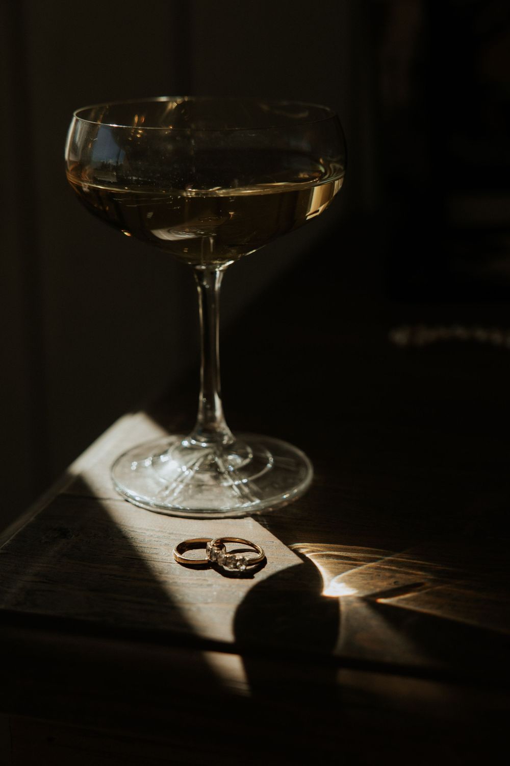 Wedding rings casting dramatic shadows beside a wine glass in moody low-key lighting on a wooden surface.