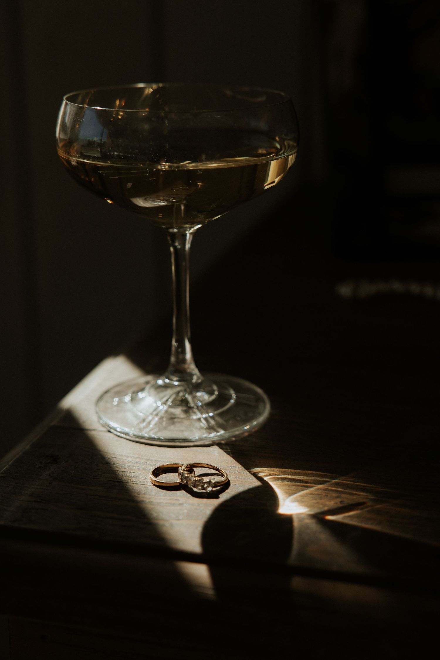 Wedding rings casting dramatic shadows beside a wine glass in moody low-key lighting on a wooden surface.