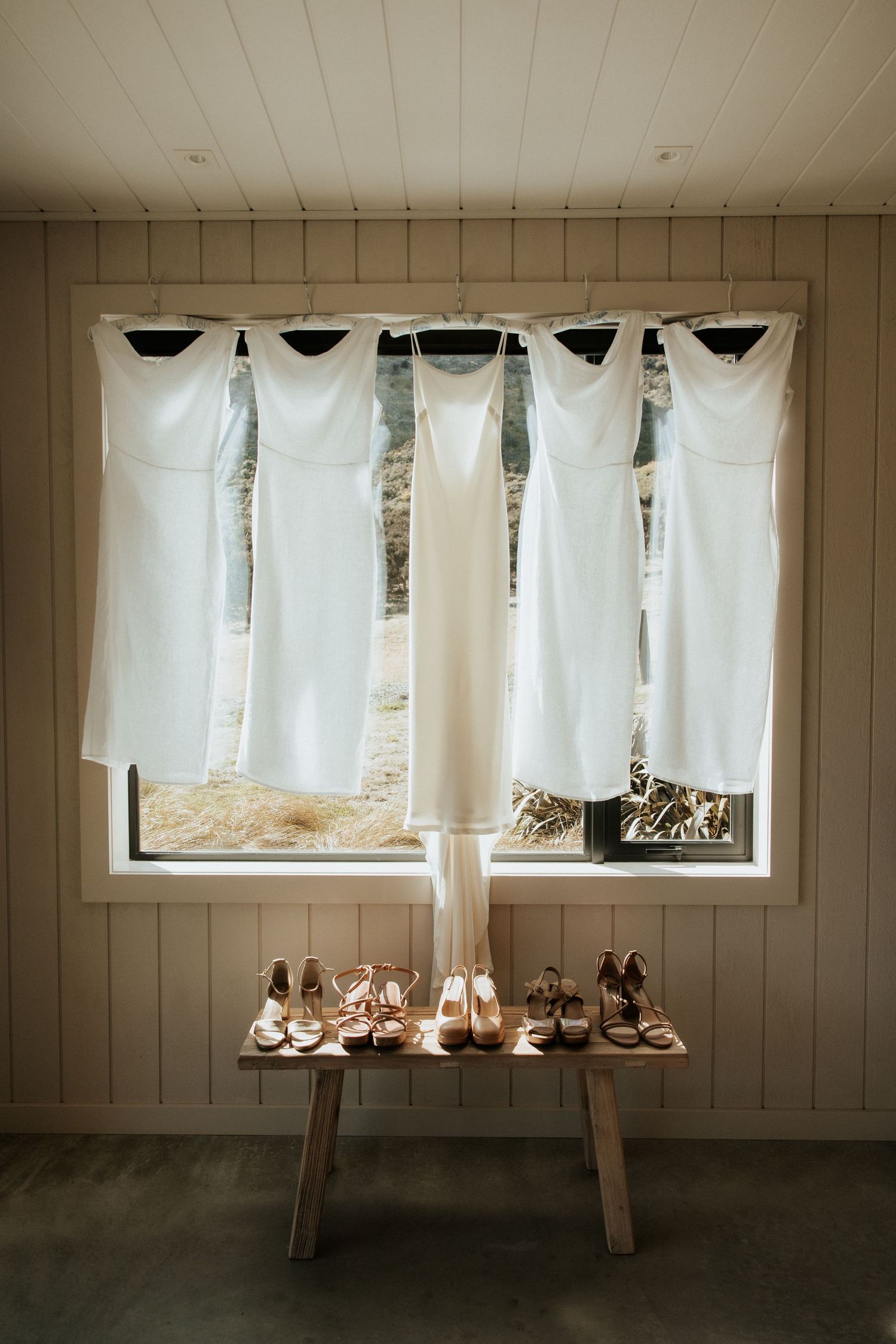 Five white bridesmaid dresses hanging in a window with matching shoes arranged on a wooden bench below.