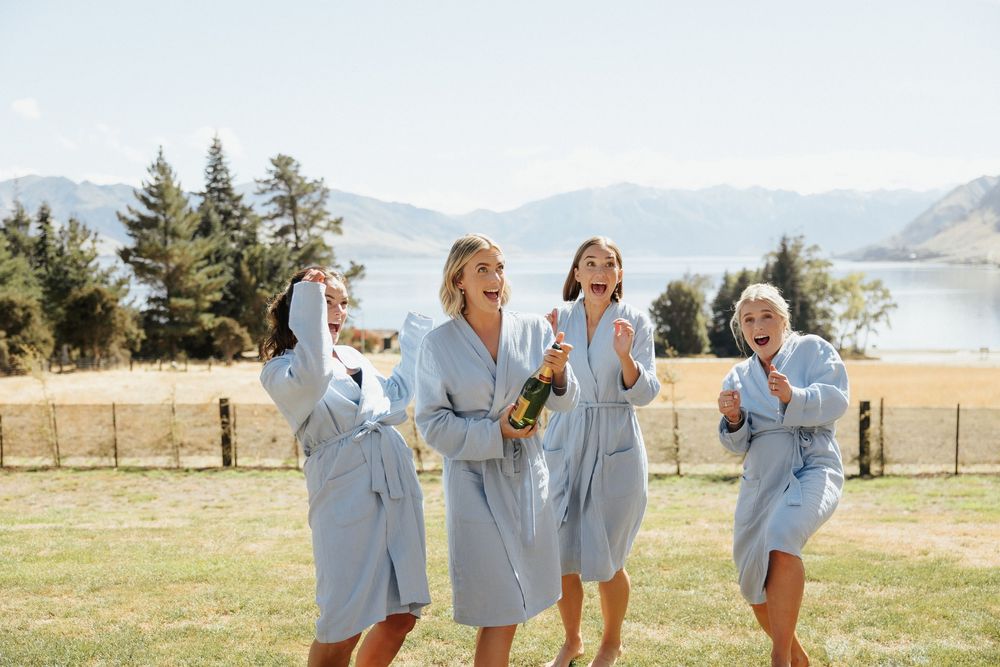 Group of women in matching light blue robes laughing and celebrating outdoors with mountains and lake in background.