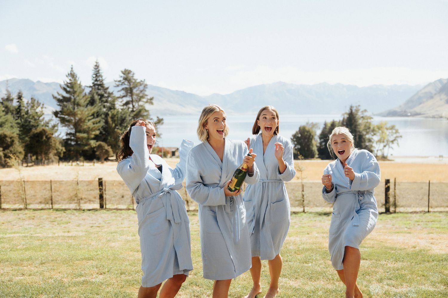 Group of women in matching light blue robes laughing and celebrating outdoors with mountains and lake in background.
