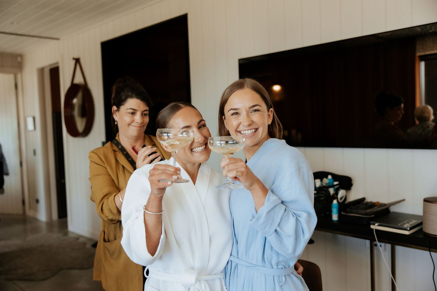 Three people smile while holding coffee mugs together in a bright home interior.
