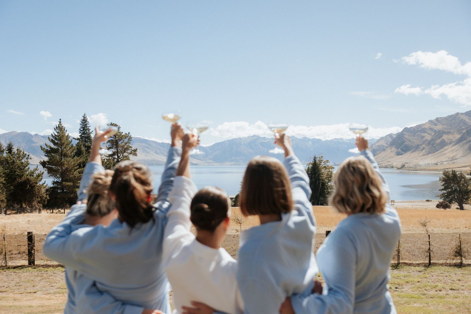 A group of friends raise their hands in celebration while sitting together overlooking a scenic mountain lake landscape.