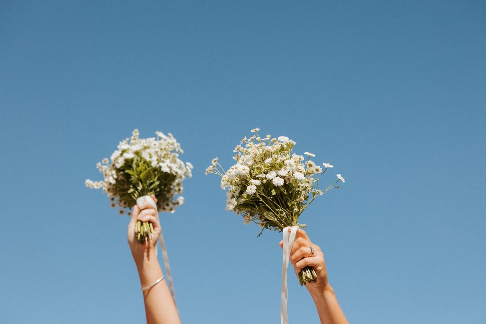 Two hands holding up white wildflower bouquets against a bright blue sky on a sunny day.