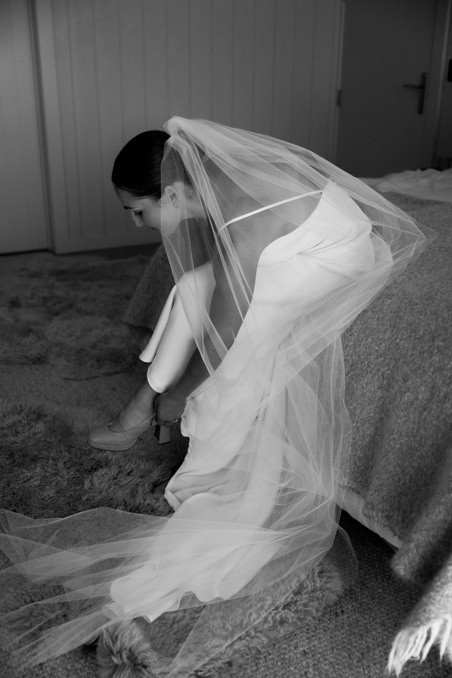 A bride in a flowing white veil sits down to put on her shoes in a black and white photograph.