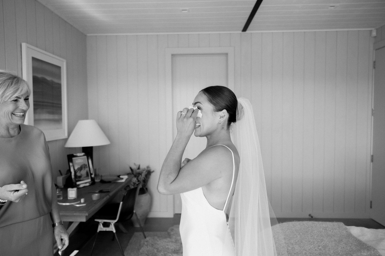 A person in a white wedding dress wipes their eye while wearing a long veil as they get ready in a bedroom.