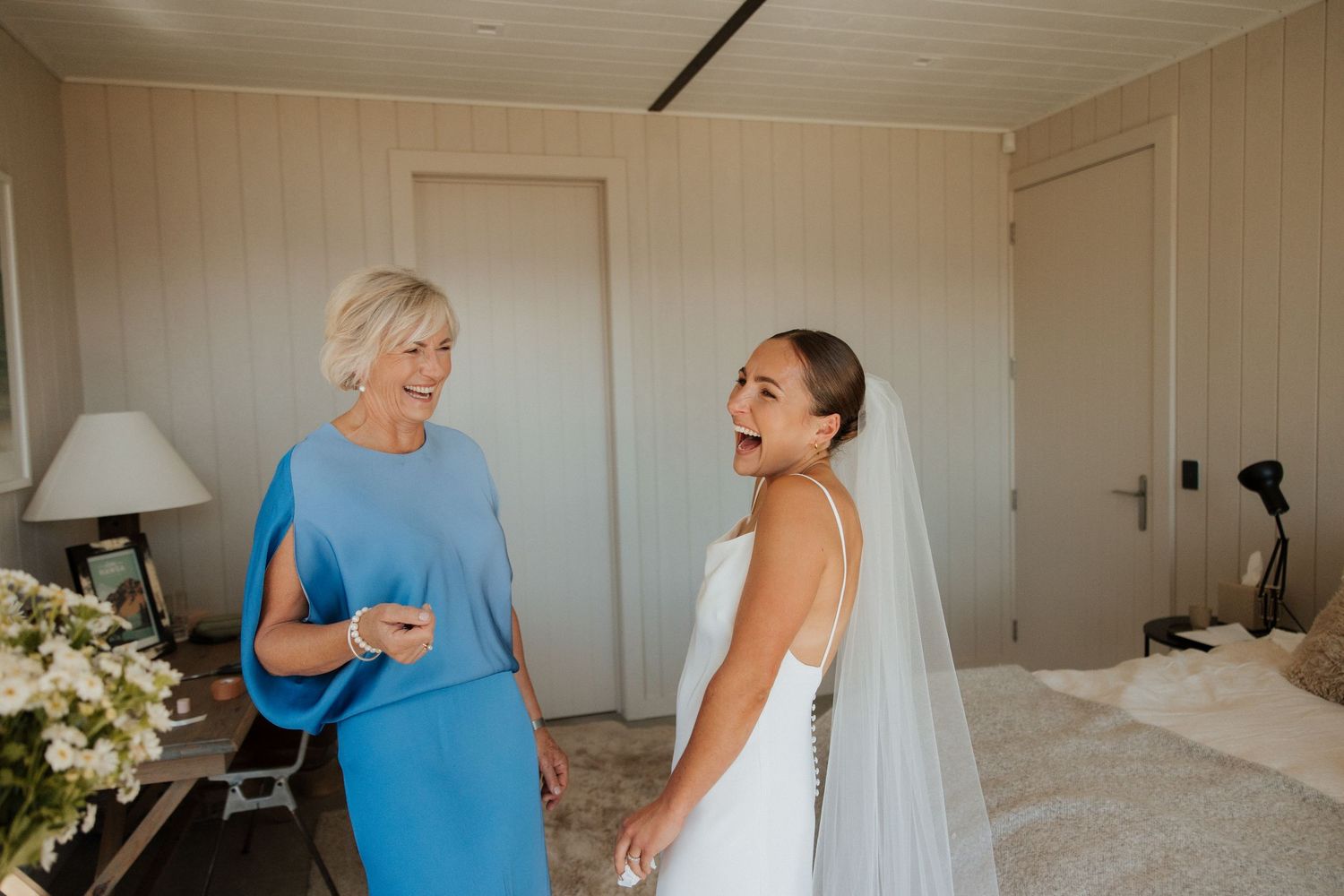 A bride in a white wedding dress and veil shares a joyful moment with a woman in a bright blue dress.