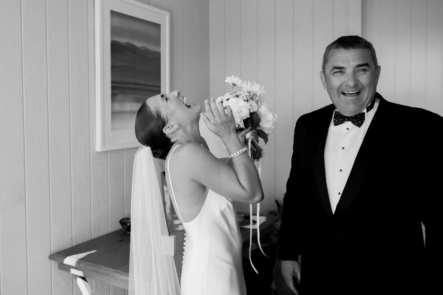 A laughing bride in a white gown dips backward while holding her bouquet next to a person in formal attire.