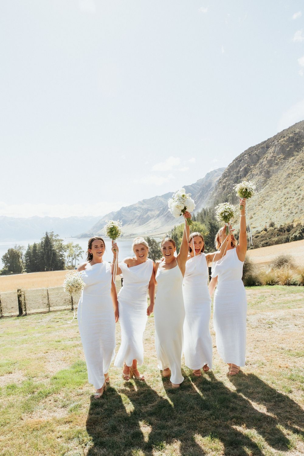 Group in flowing white dresses holding bouquets aloft against mountainous backdrop on sunny day.