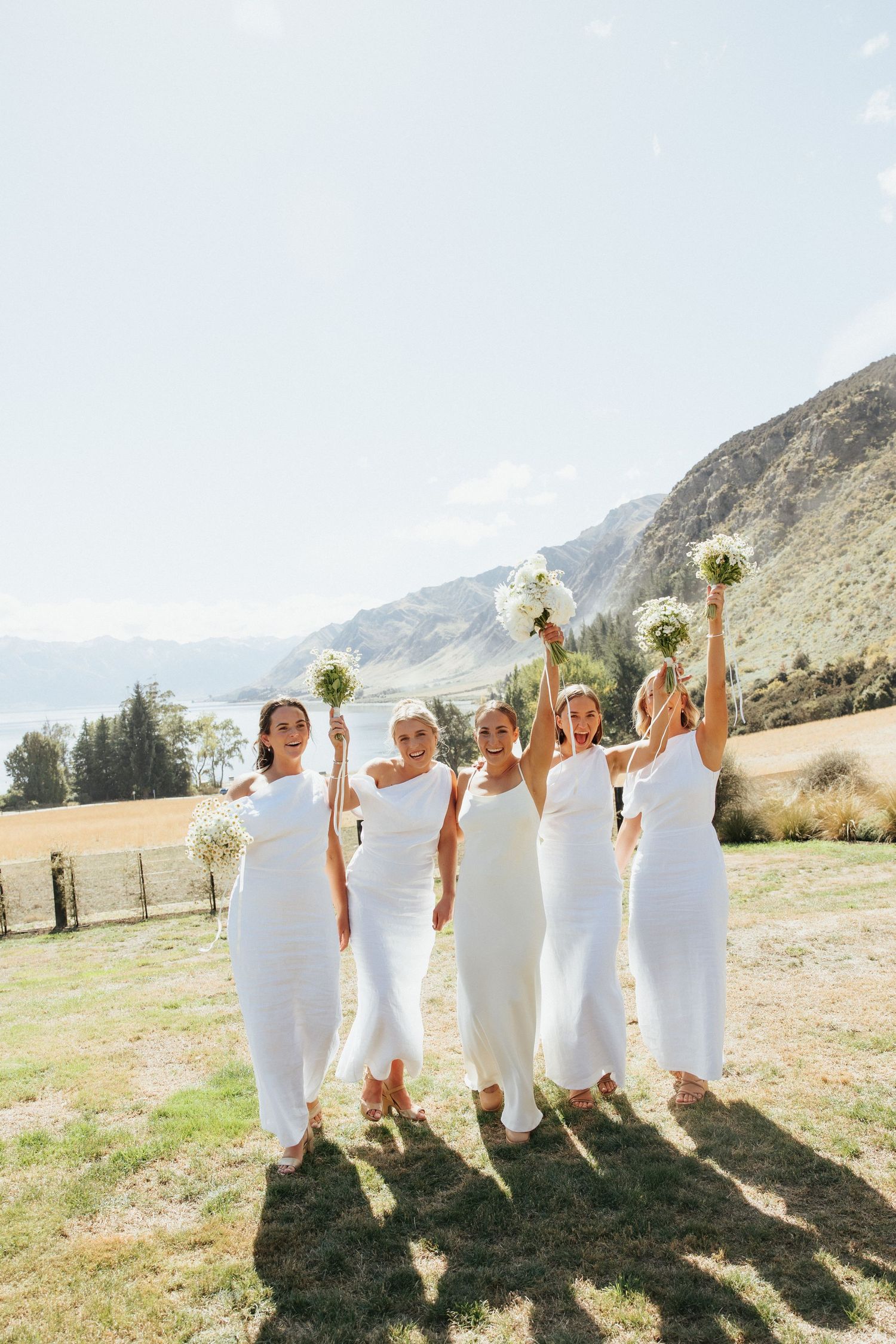 Group in flowing white dresses holding bouquets aloft against mountainous backdrop on sunny day.