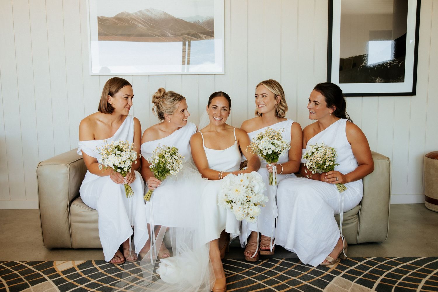 Bridal party in elegant white dresses sit together on beige couch holding beautiful floral bouquets.