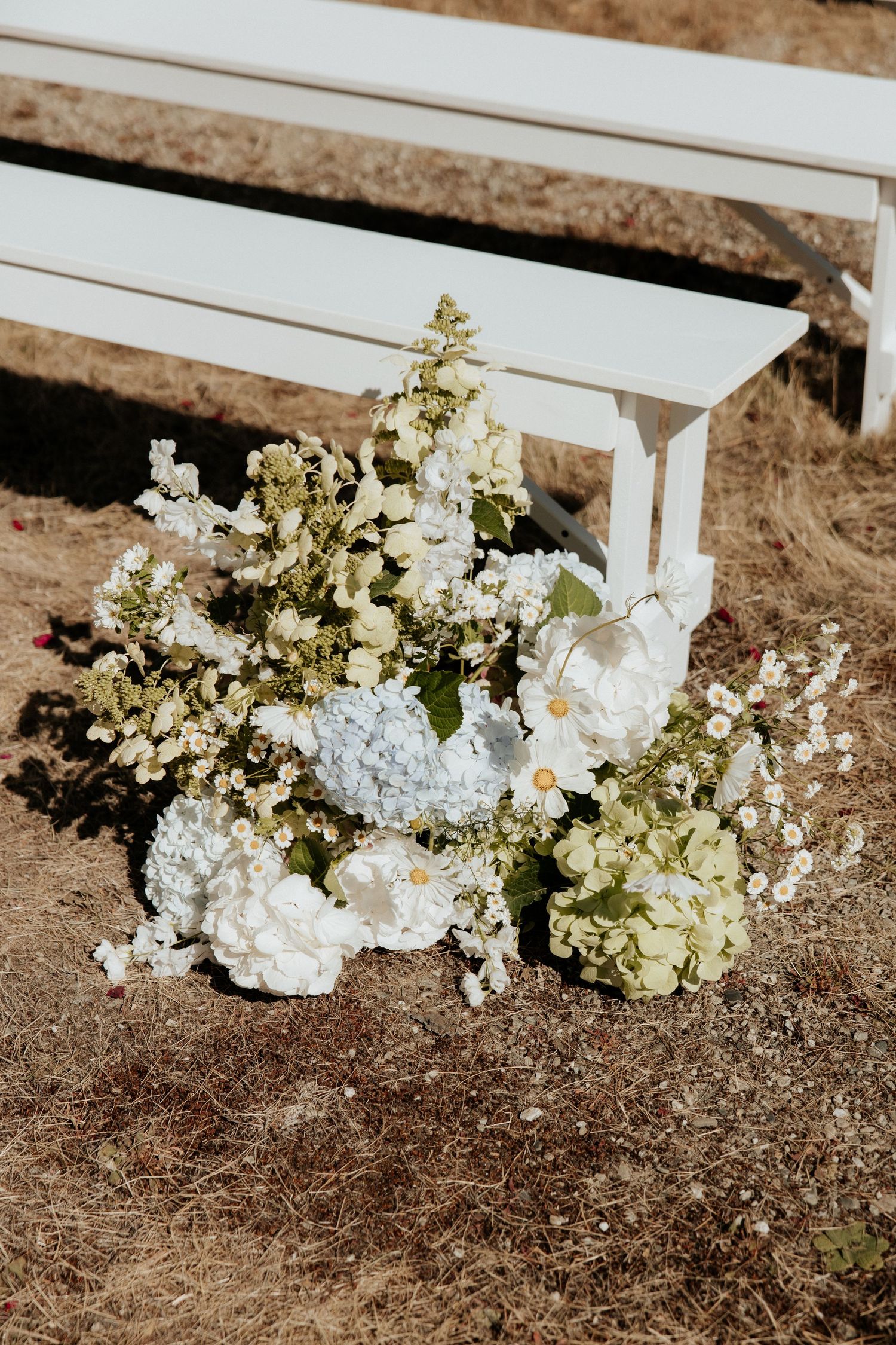 Elegant white and cream floral arrangement with hydrangeas and greenery against a rustic bench backdrop.