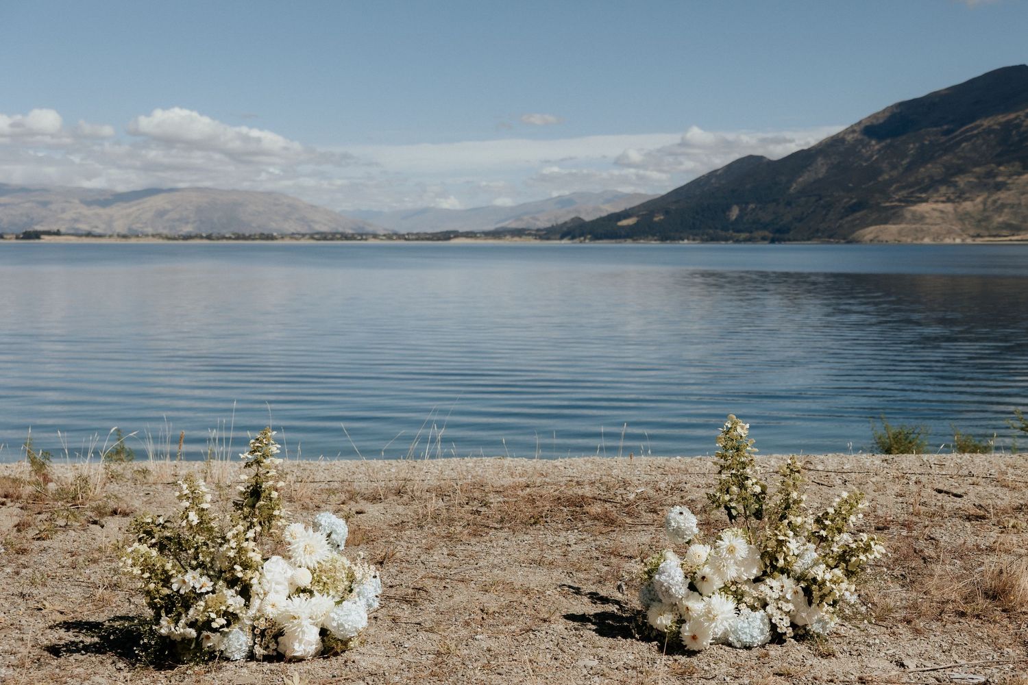 White floral arrangements decorate a lakeside beach with mountains in the background on a sunny day.