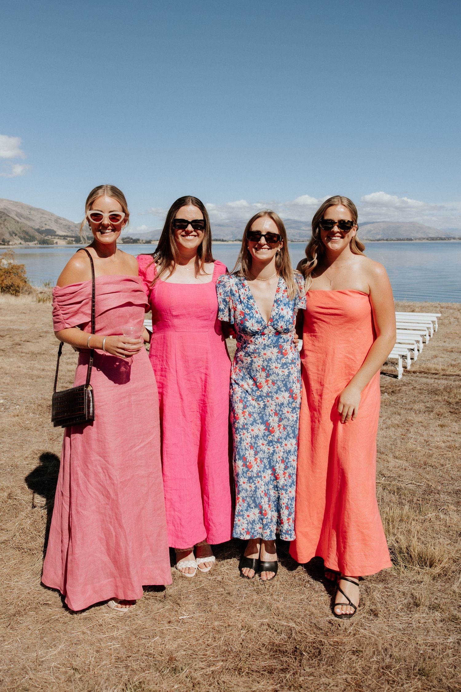 Bridesmaids in coral and pink dresses pose together against a scenic mountainous backdrop near a lake.