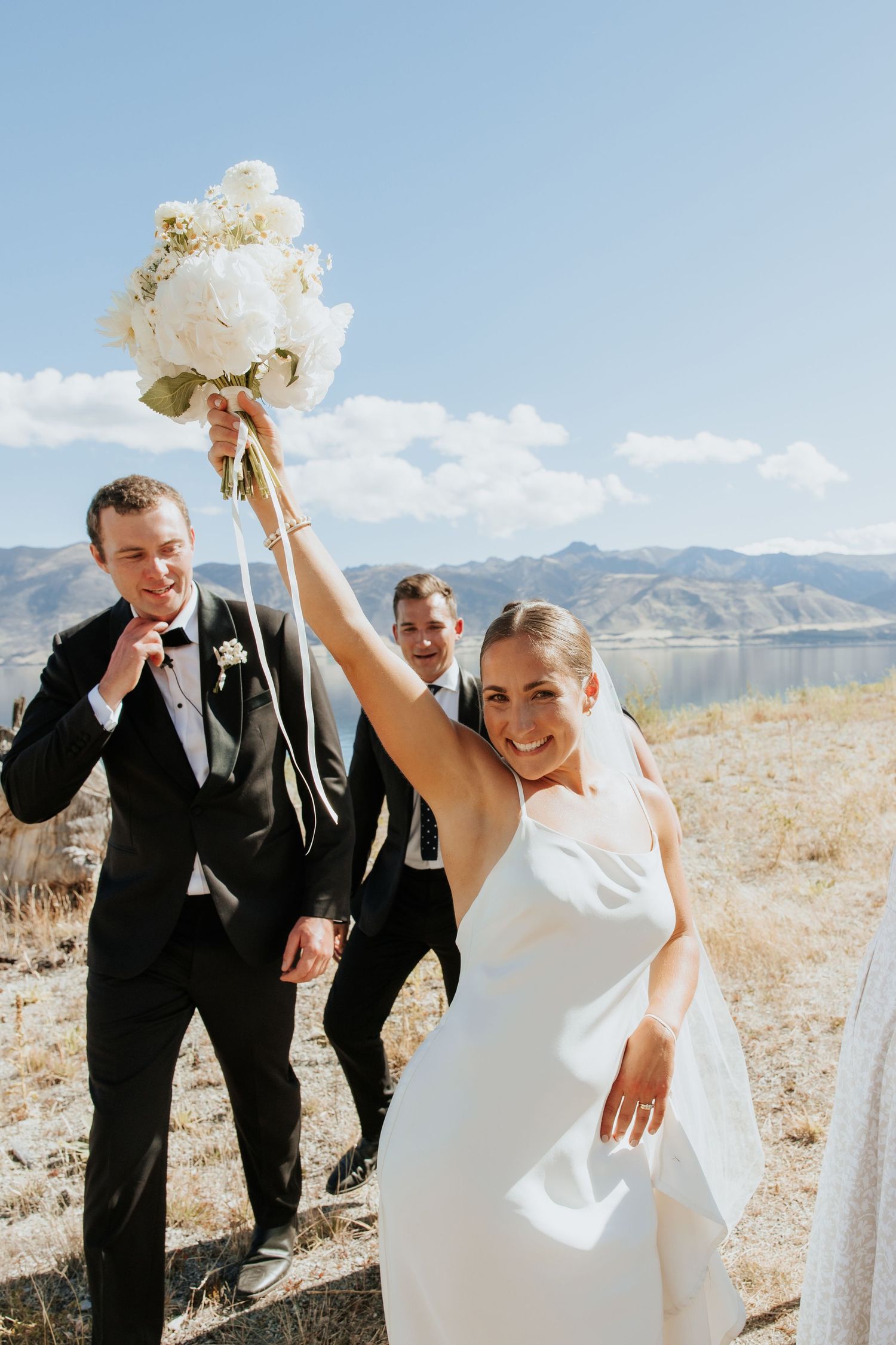 Wedding party celebrating with raised bouquet against mountain lake backdrop on sunny day.