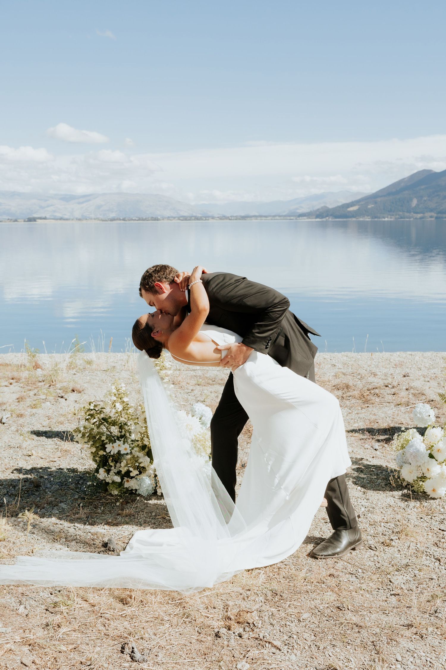A romantic wedding couple share an intimate dip kiss against a scenic mountain lake backdrop.