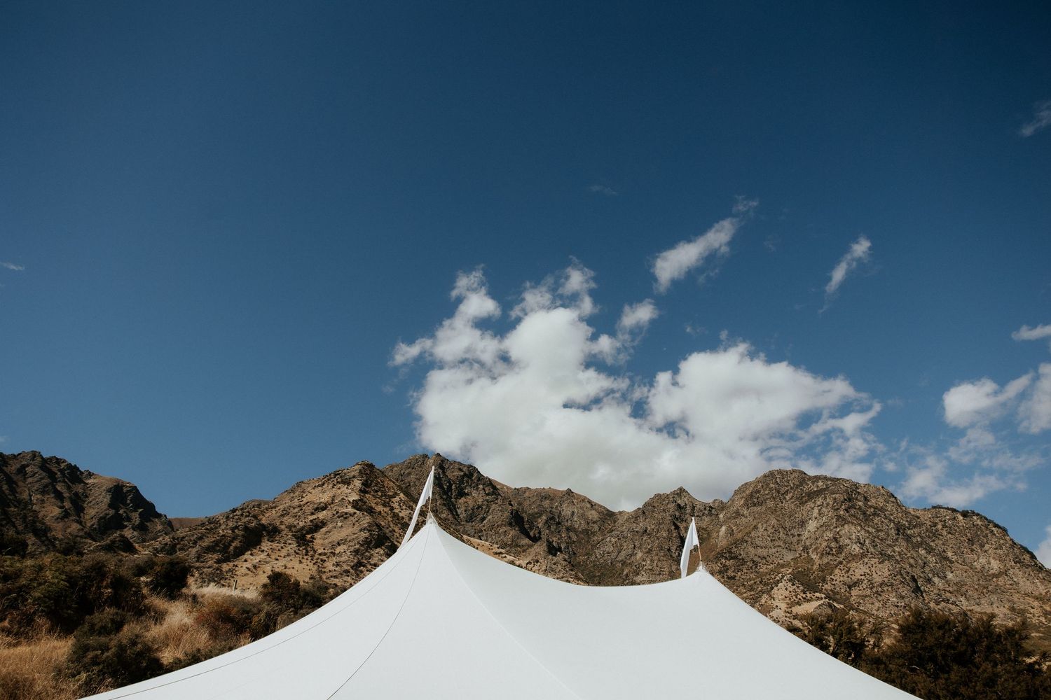 A white wedding tent peaks against a rugged mountain landscape under bright blue skies.