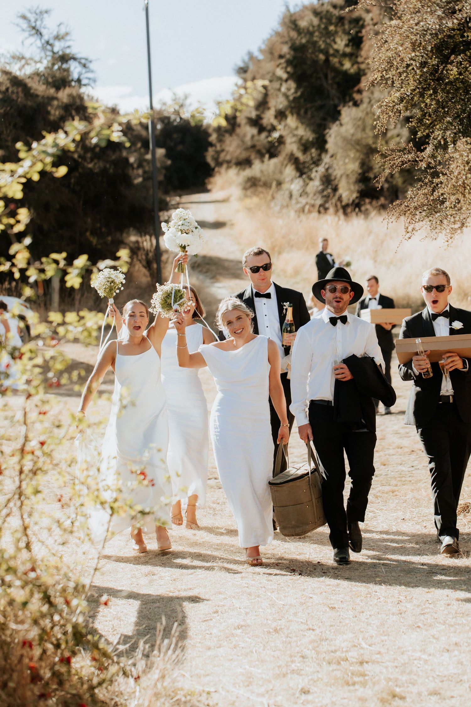 Wedding party walks joyfully down a sunlit path surrounded by blooming trees.