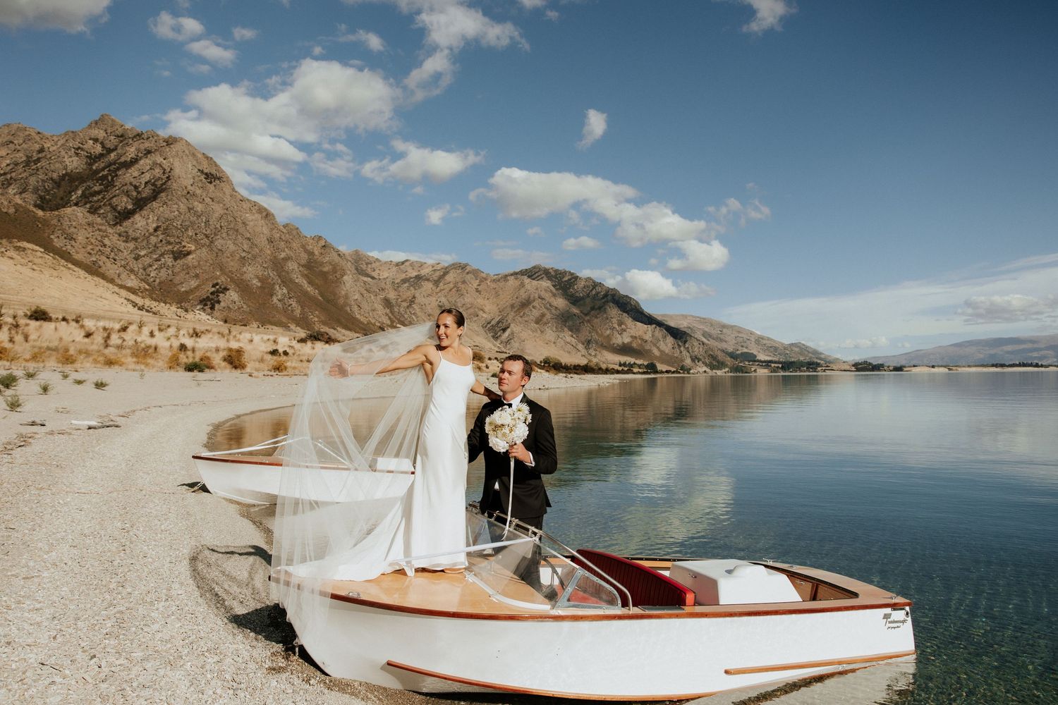 A couple poses on a vintage wooden boat by a serene mountain lake on their wedding day.