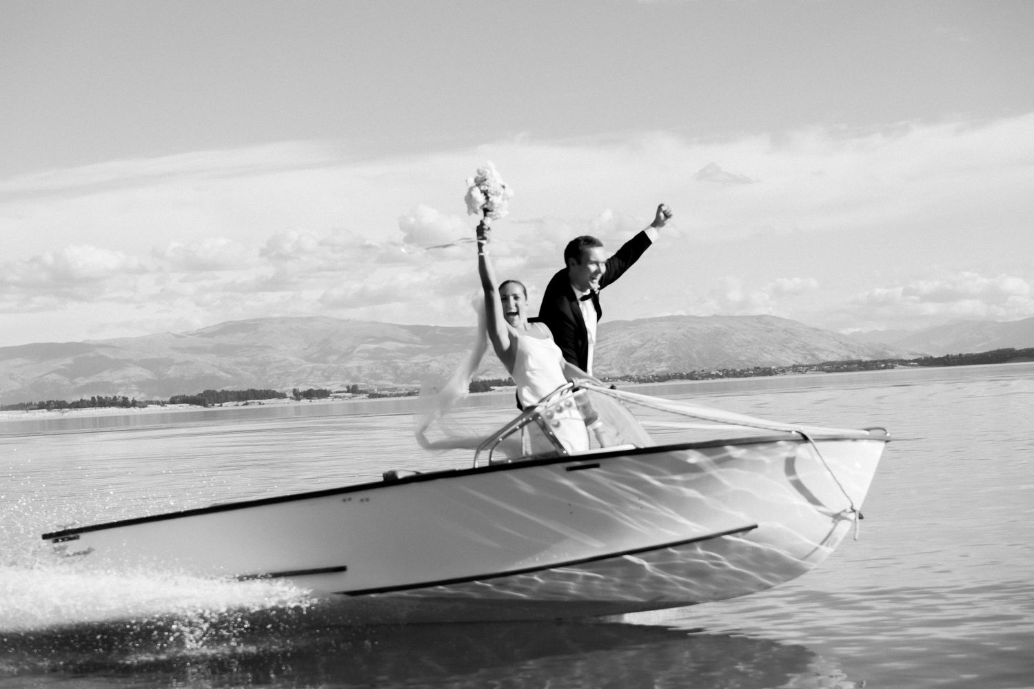 Two figures joyfully pose on a boat gliding across a lake with mountains in the background.