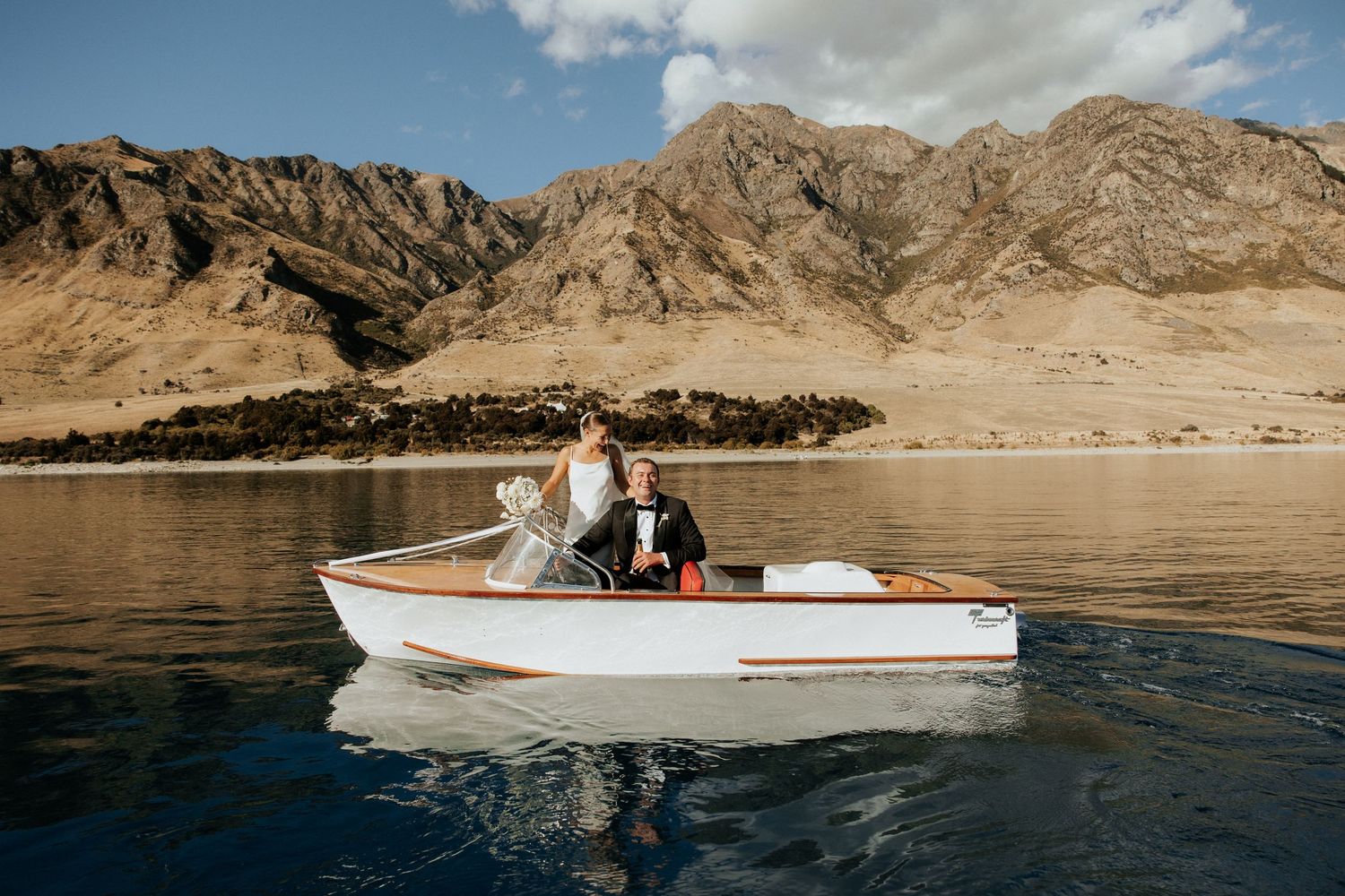 Newlyweds enjoying a romantic boat ride on a lake surrounded by majestic mountains.