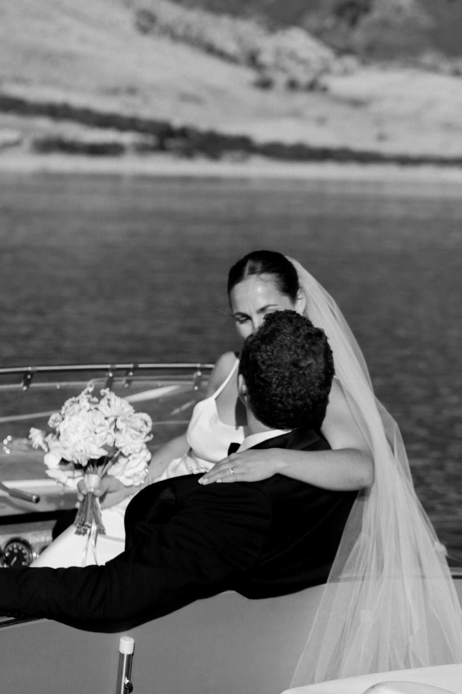 Newlywed couple sitting together on a boat with water and mountains in the background.