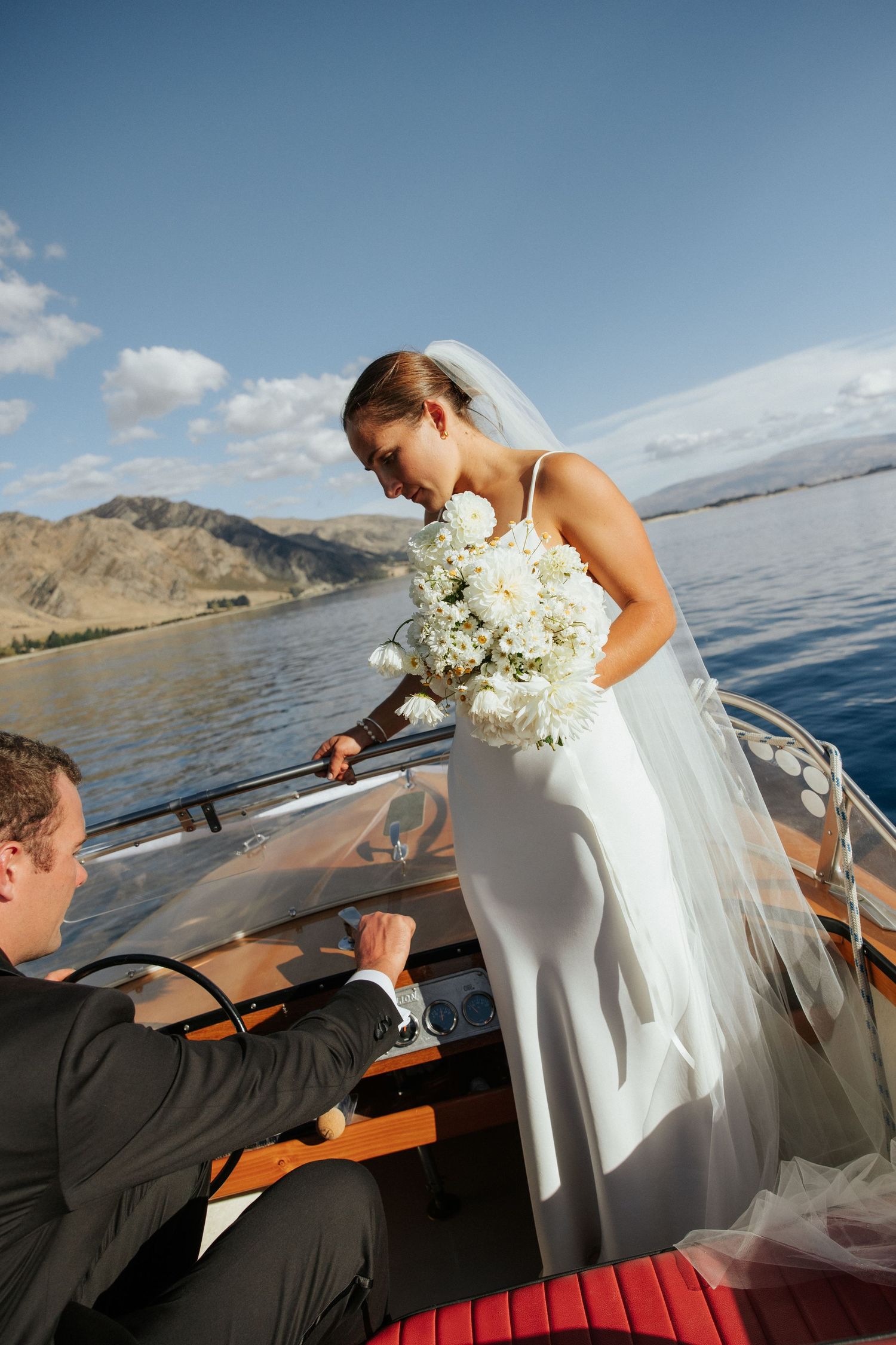White-clad figure holding bouquet on speedboat against mountain backdrop.