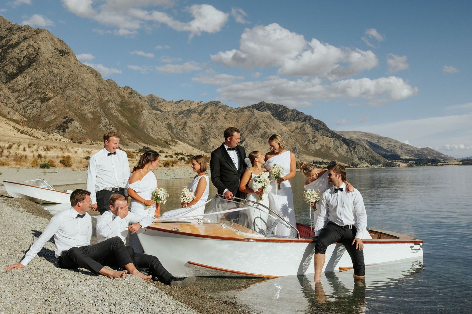 Wedding party poses casually on wooden boat by scenic mountain shoreline.