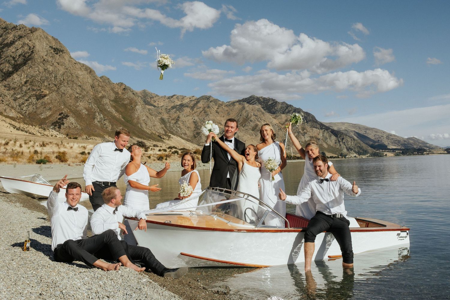 Wedding party posing playfully on vintage boat by rocky mountain shoreline.