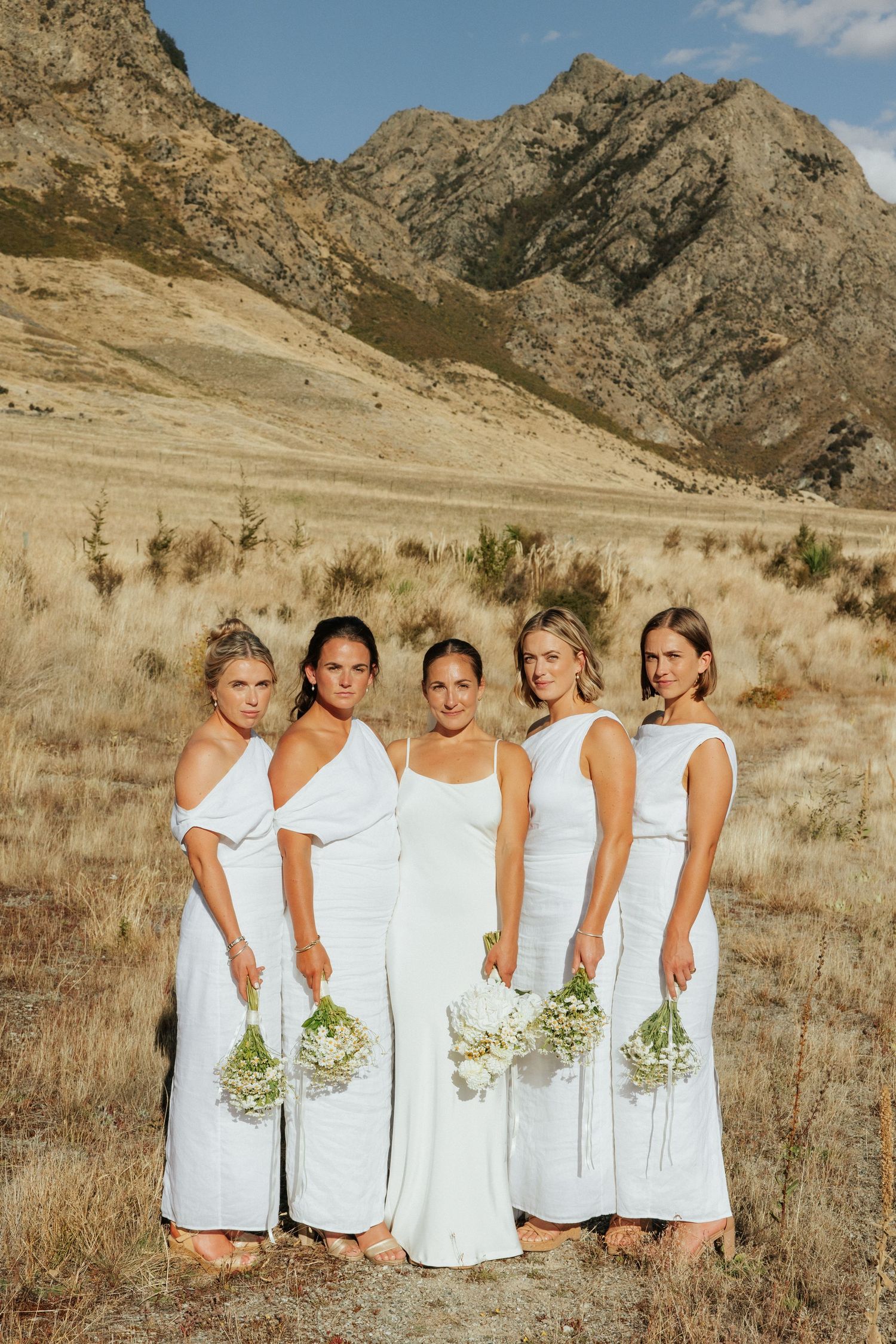 Bridesmaids in white dresses holding bouquets pose against mountain landscape backdrop.