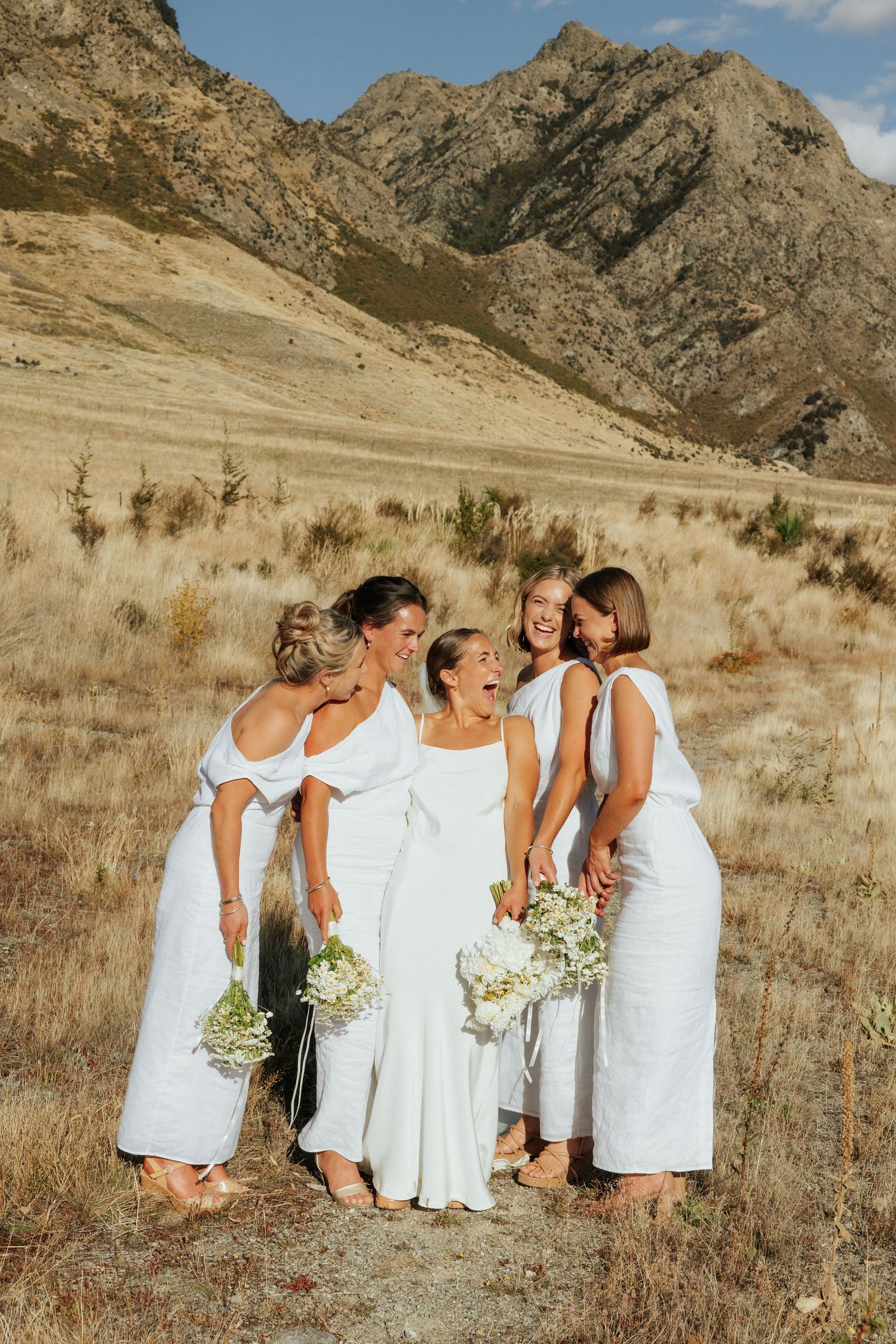 Group in white dresses stands together holding bouquets against rugged mountain landscape on sunny day.