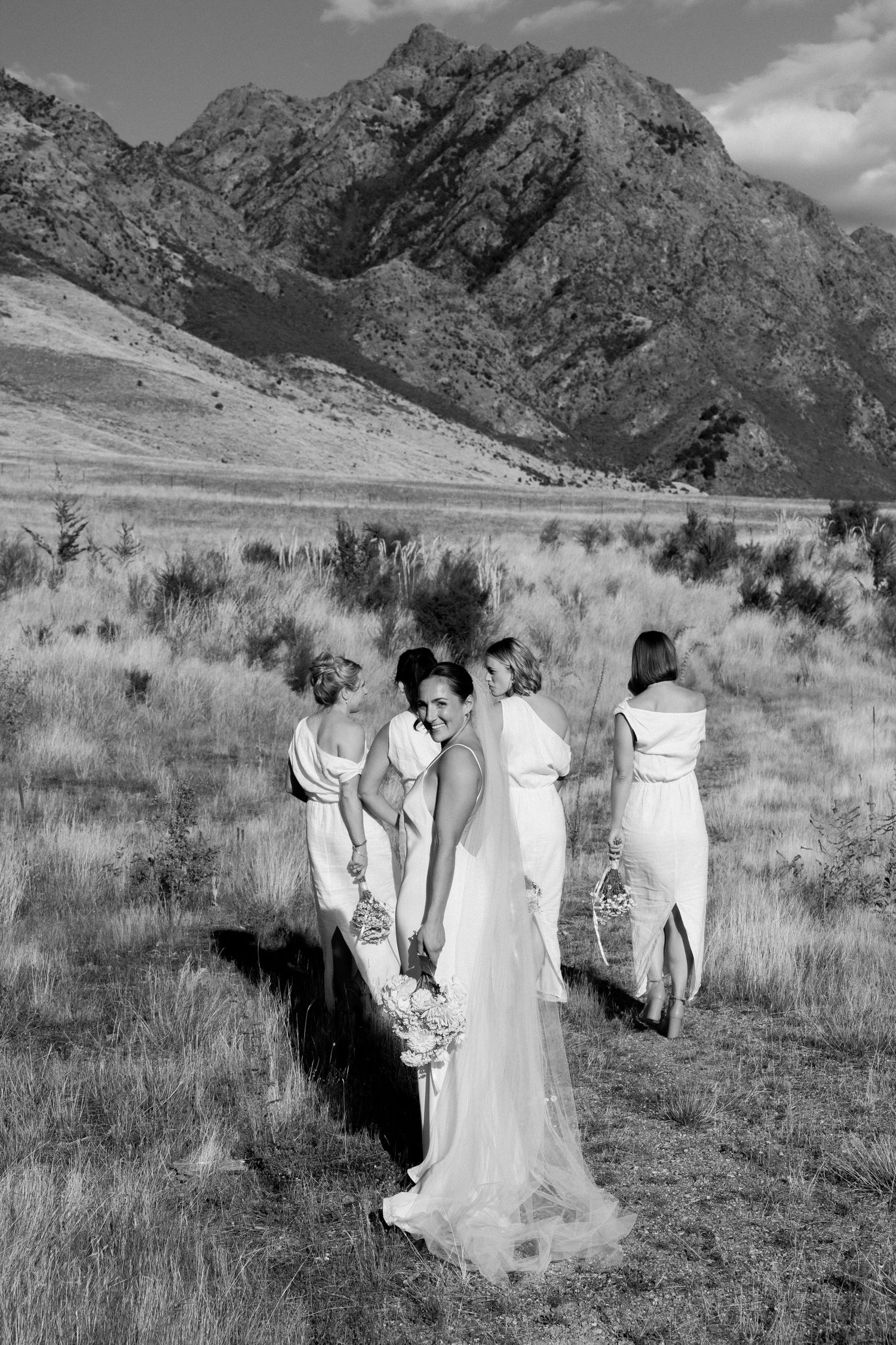 Group in elegant white dresses posing in field with dramatic mountain landscape.