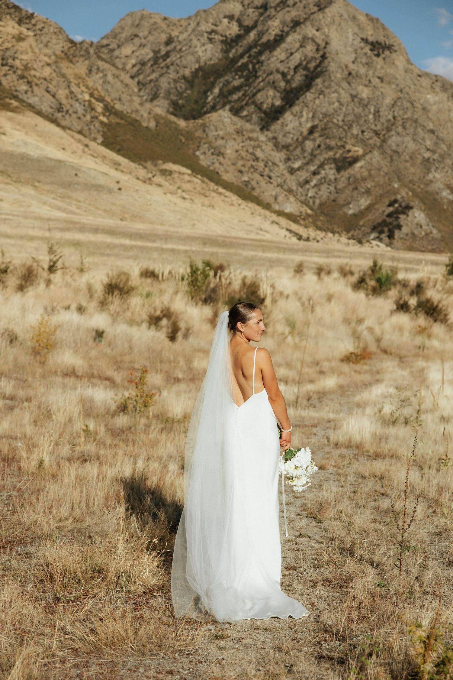 Elegant figure in white wedding dress standing in mountain field holding bouquet.