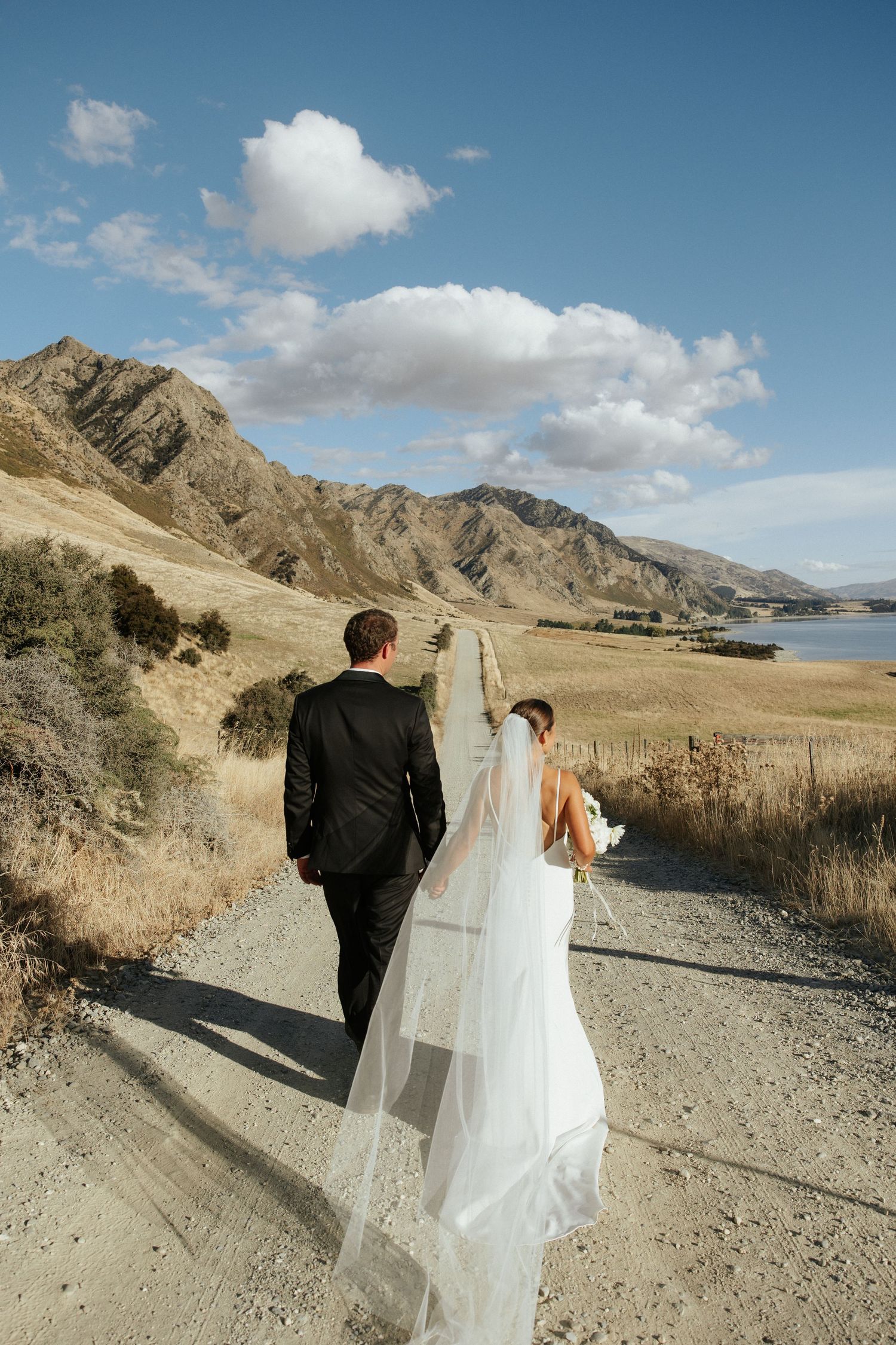 A couple on a scenic mountain path with a flowing white wedding dress and lake views in the background.