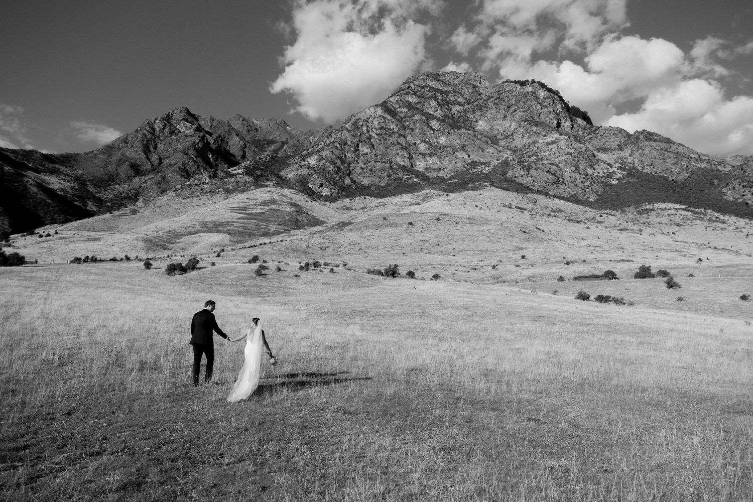 Black and white photo of couple walking across mountain meadow with dramatic peaks in background.