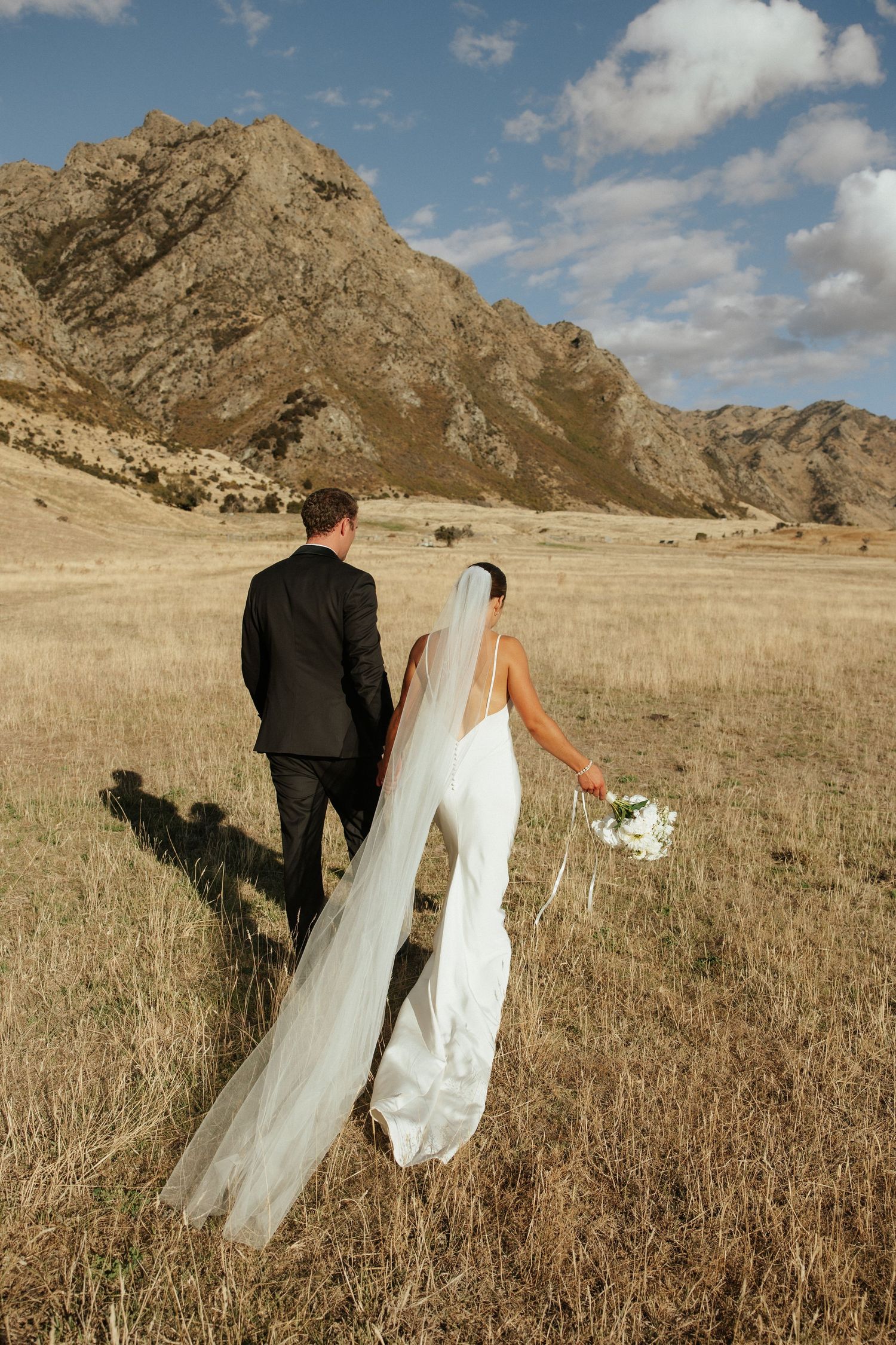 A couple in wedding attire stands together in a golden field with dramatic mountain peaks rising in the background.