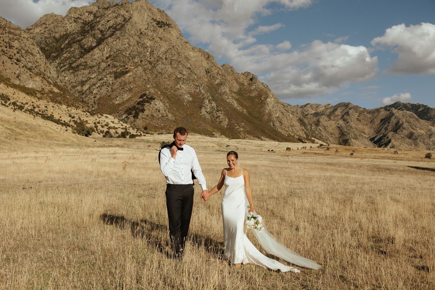 A newlywed couple holds hands while walking through a golden field with majestic mountains behind them.