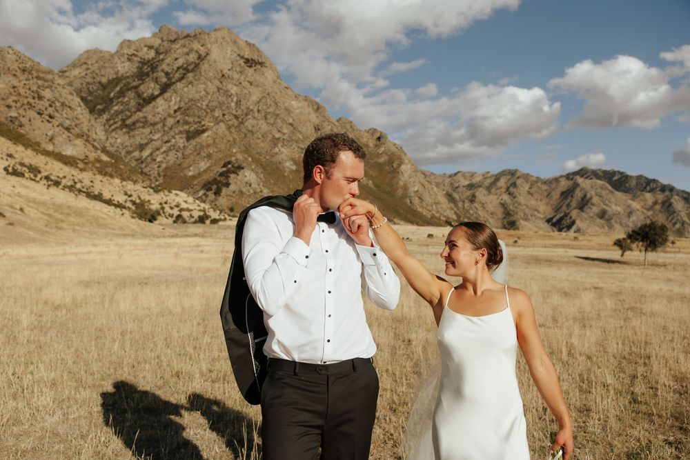 Newlyweds share a playful moment in golden field with mountains behind.