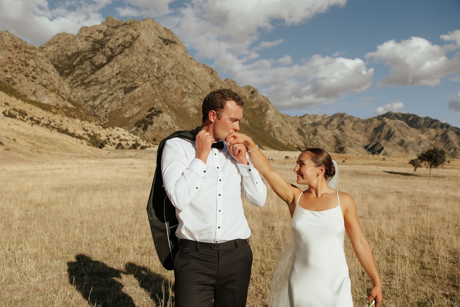 Newlyweds share a playful moment in golden field with mountains behind.