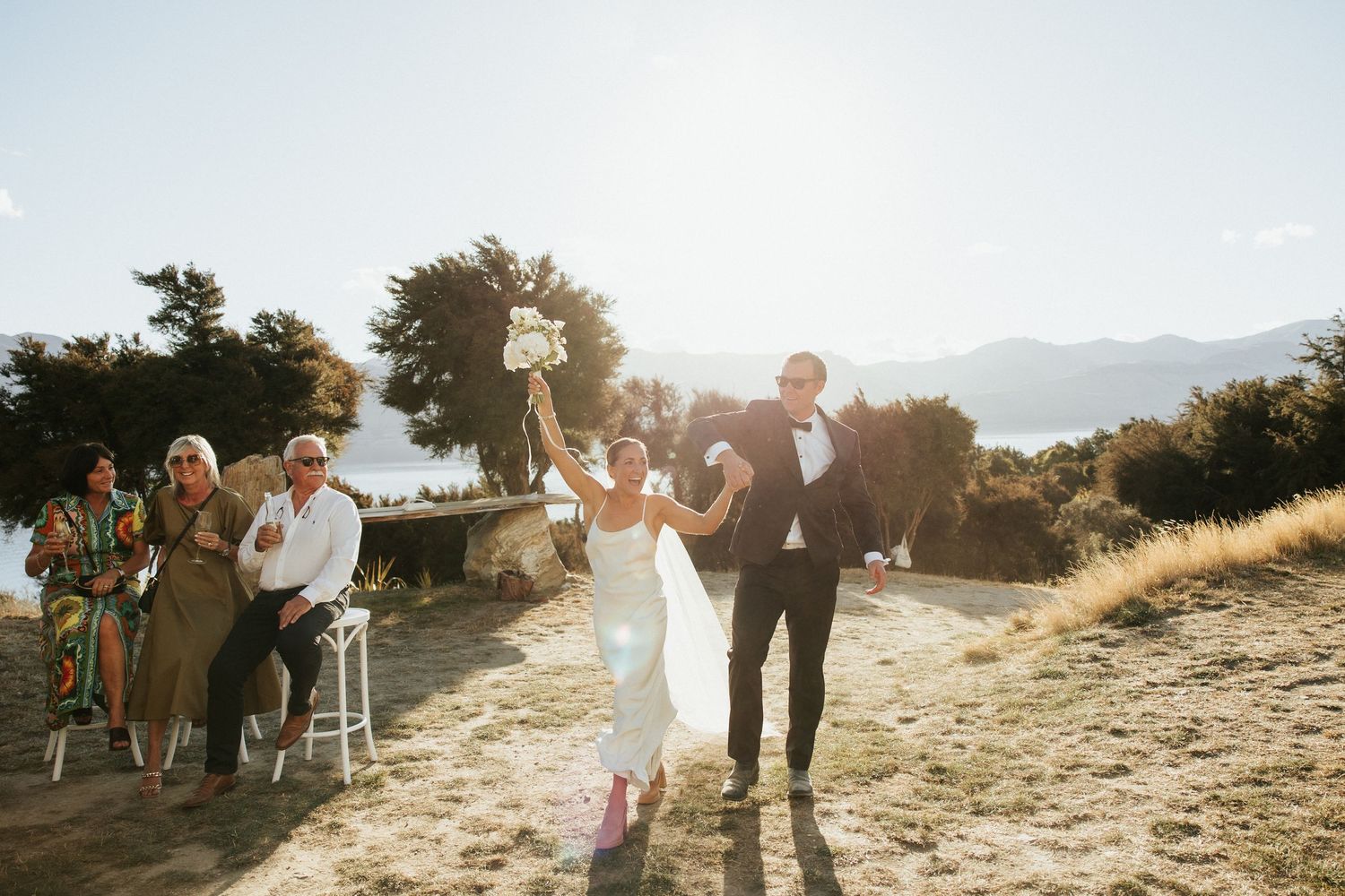 A wedding party celebrates on a hilltop path at sunset with mountains in the distance.