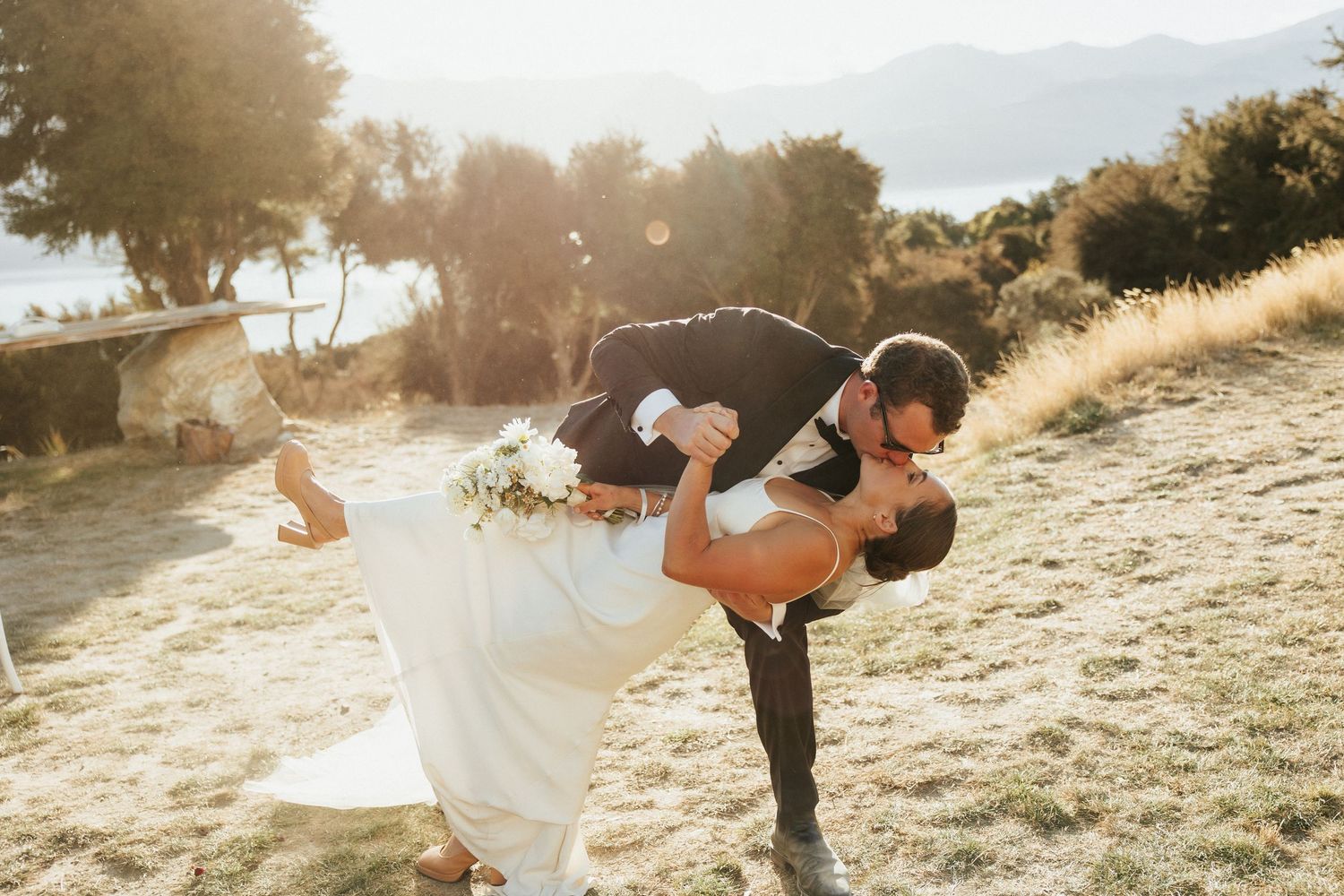 Romantic sunset dip kiss between newlyweds on mountainside with glowing backlight.