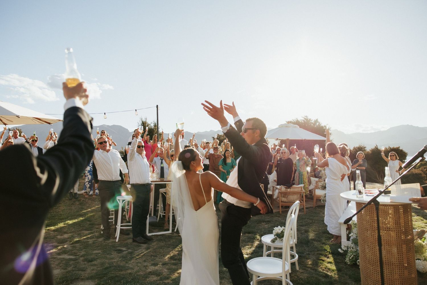 Wedding celebration outdoors with guests cheering as newlyweds dance under a mountain landscape.