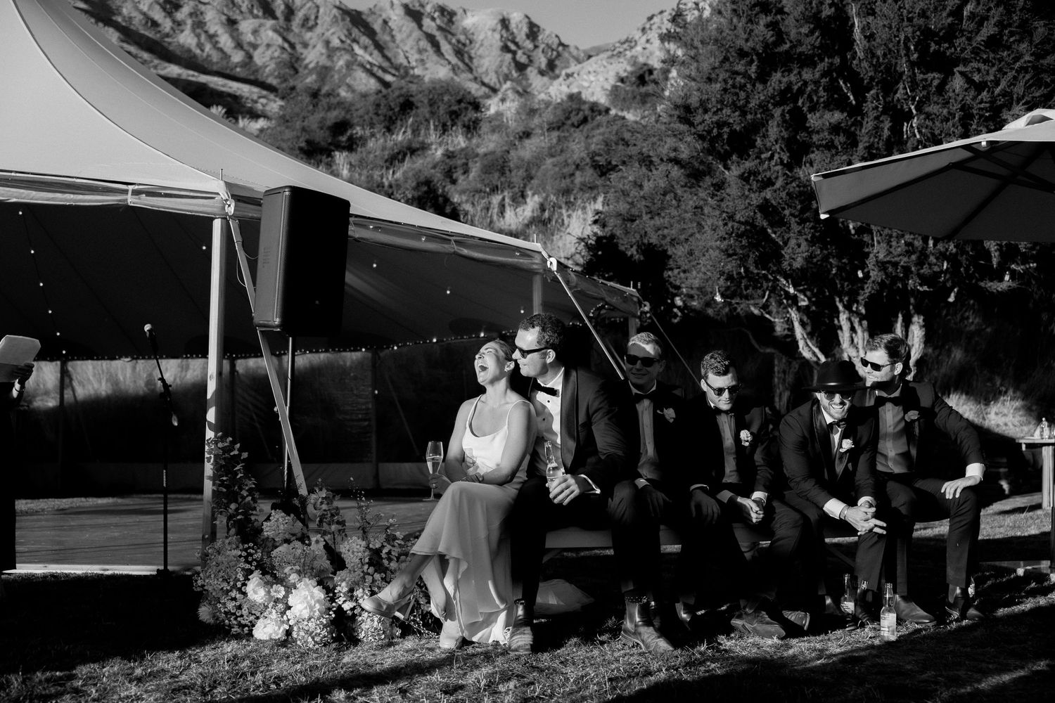 Black and white photo of wedding party seated under a tent during outdoor celebration.