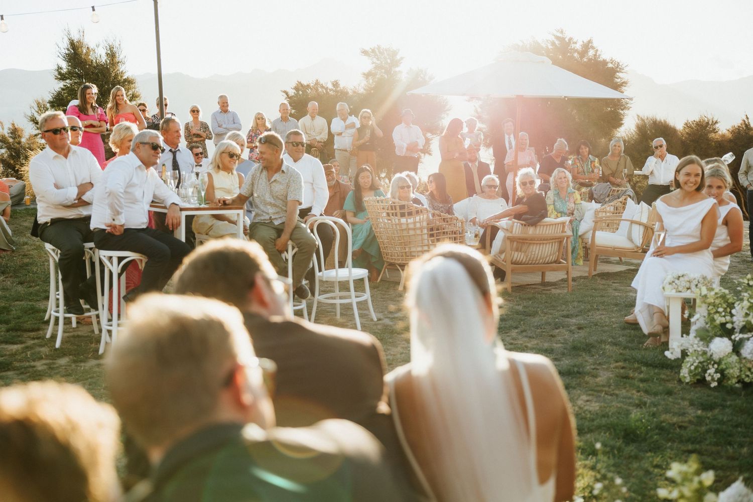 Guests gathered on white chairs at an outdoor wedding ceremony during golden hour in a garden setting.