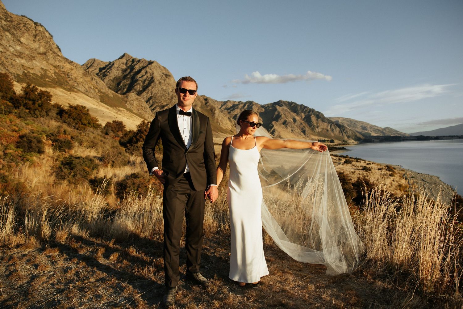 Bride and groom pose together on mountainside with flowing veil and lake view in background.