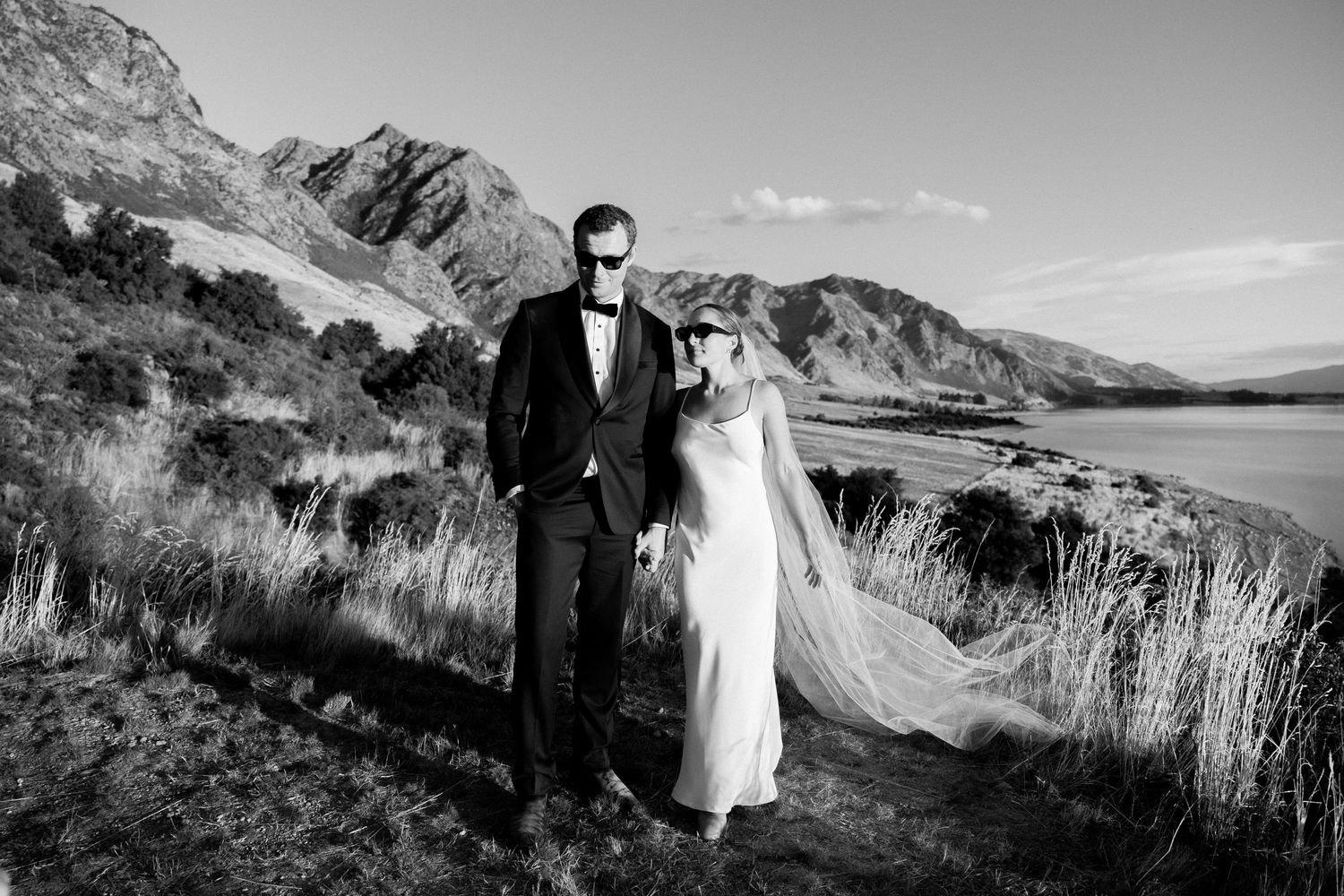 A couple poses on a scenic mountain ridge overlooking a lake in dramatic black and white photography.