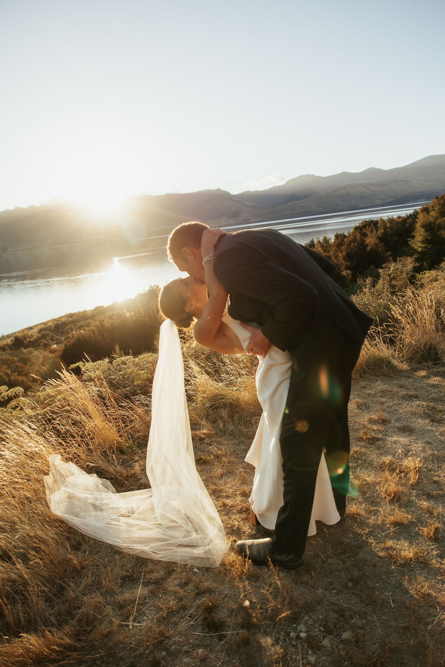 Romantic sunset moment between newlyweds with mountain lake backdrop and sunflare.